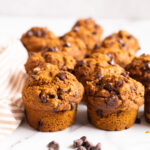 12 pumpkin chocolate chip muffins and a few chocolate chips around them with linen towel on a countertop.