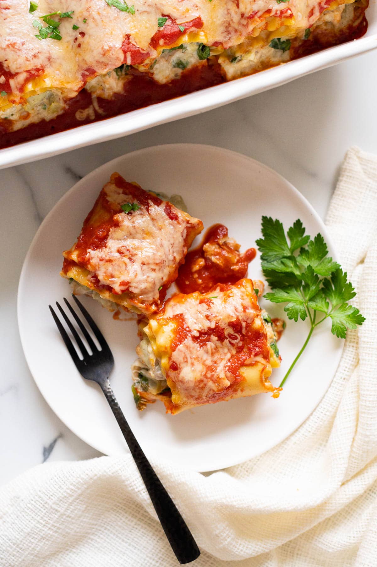 Two chicken lasagna roll-ups with sauce on a plate and parsley. More lasagna rolls in a baking dish.