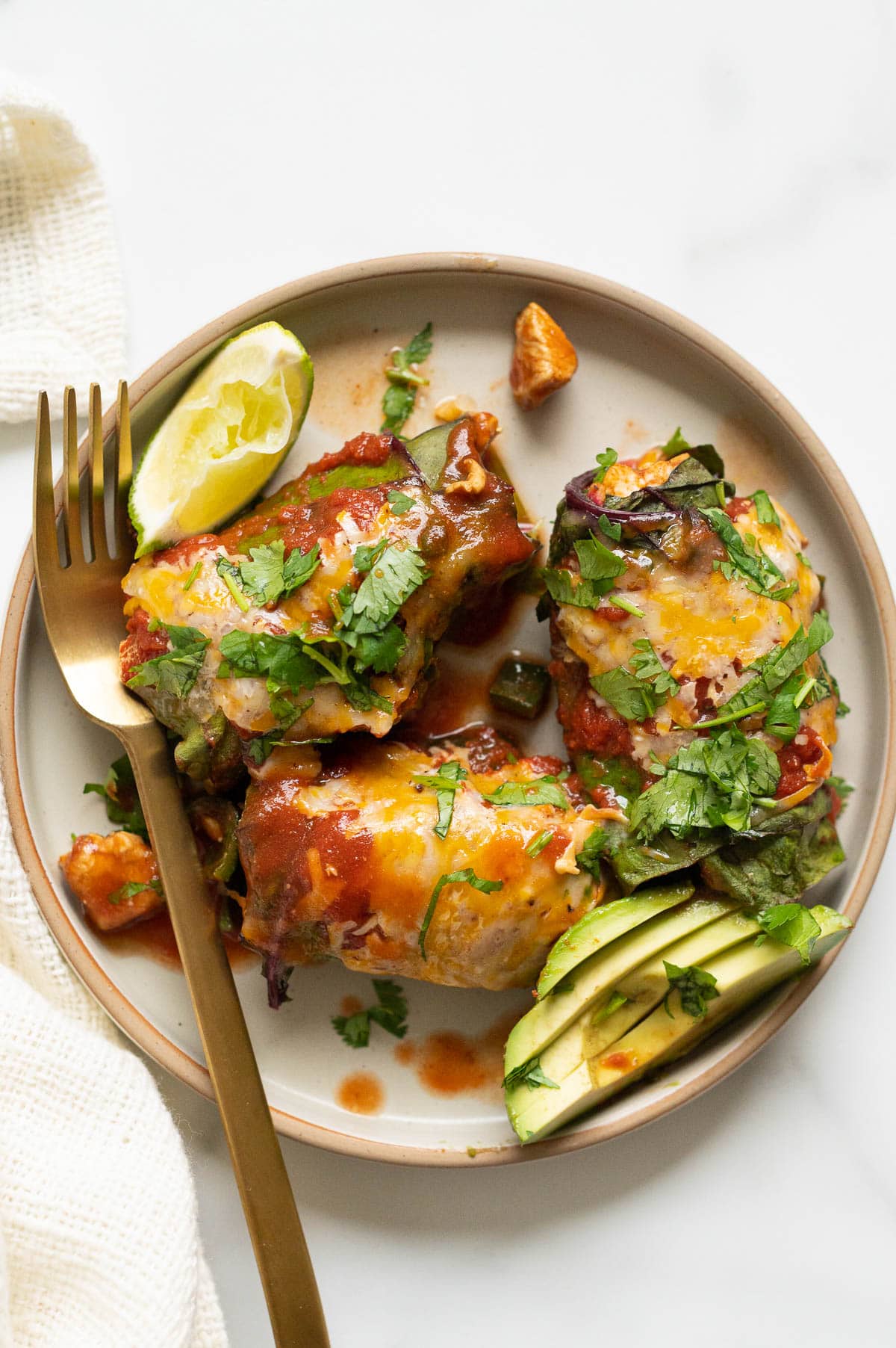 Three low carb enchiladas served with avocado, cilantro and lime on a plate with a fork.