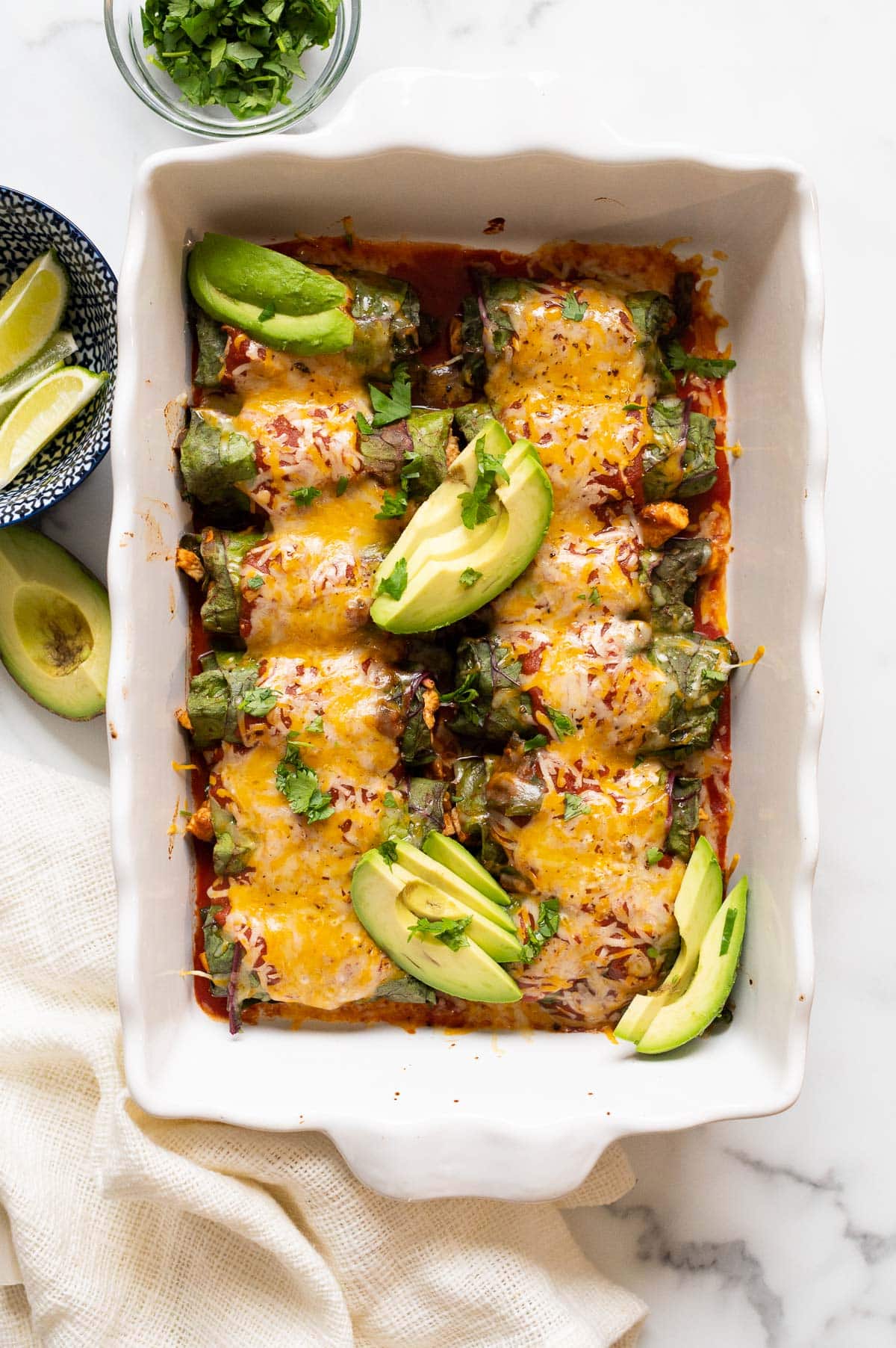 Low carb chicken enchiladas with cheese on top served in a baking dish with slices of avocado and cilantro. More avocado and lime on a side.