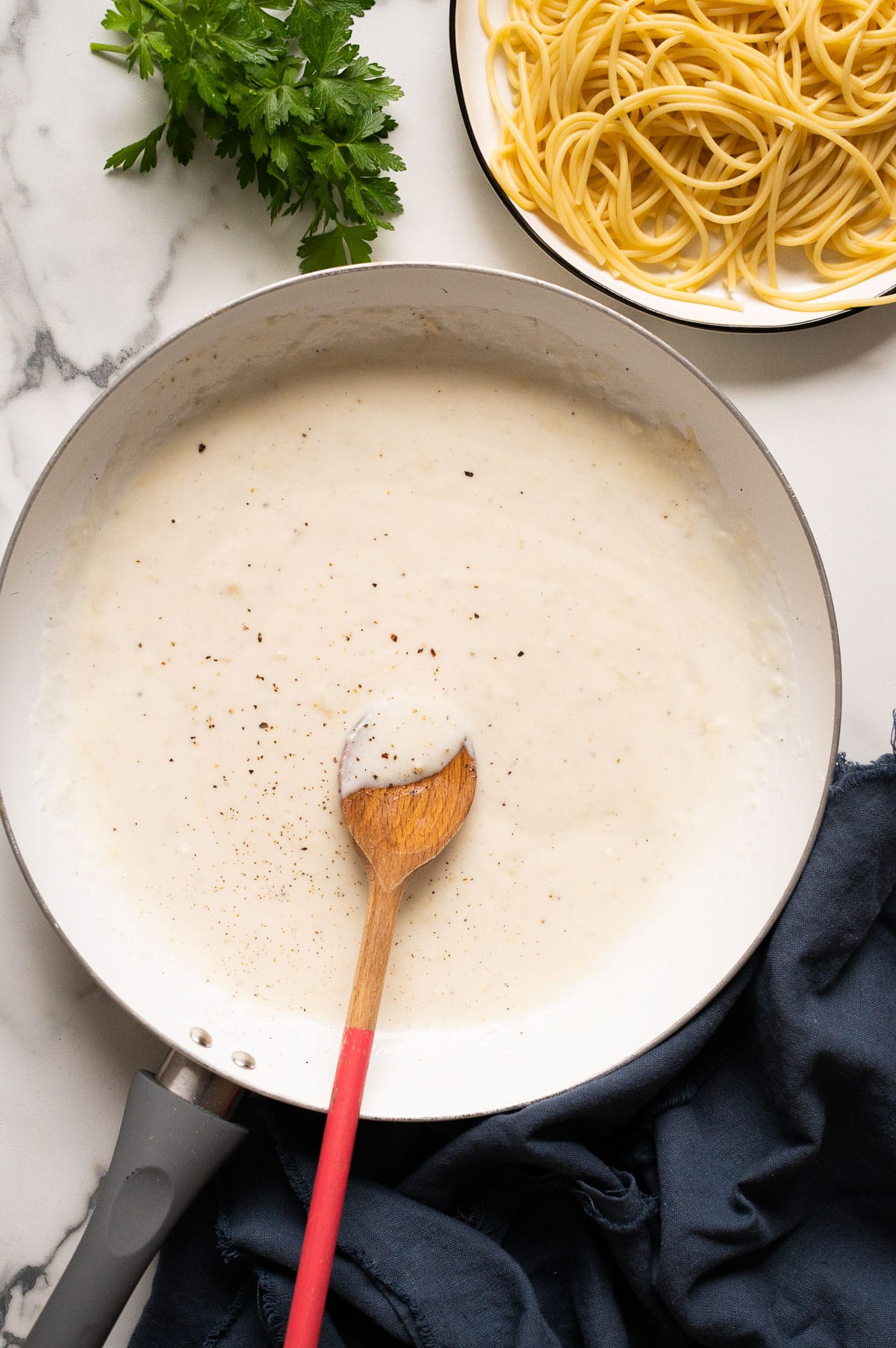 Low calorie alfredo sauce in a skillet with wooden spoon. Spaghetti on a plate, parsley and blue napkin on a countertop.