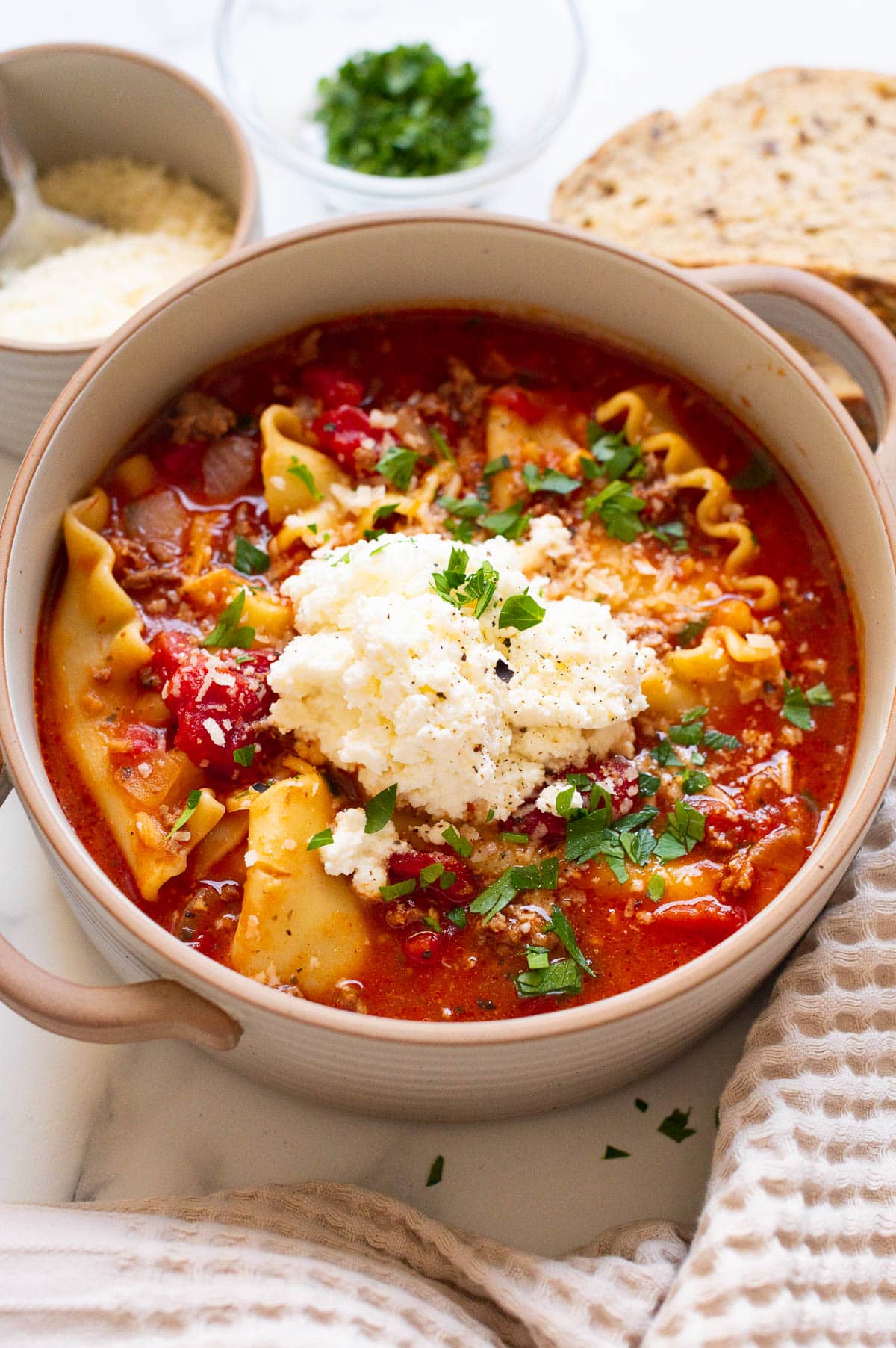 Lasagna soup with a dollop of ricotta cheese and parsley in a bowl. Bread, parmesan cheese and parsley on a side.