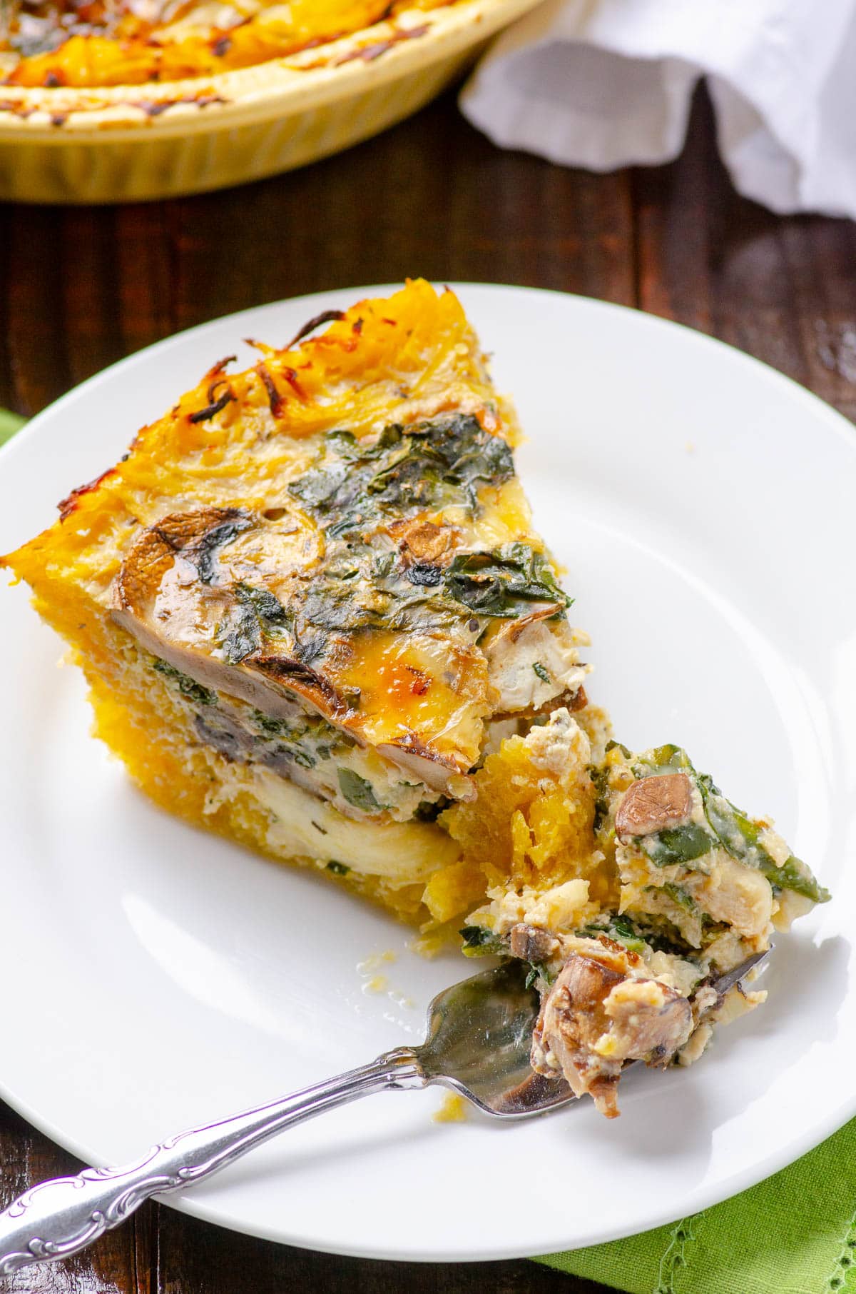 A slice of spaghetti squash quiche with kale and mushrooms with a fork on a plate.