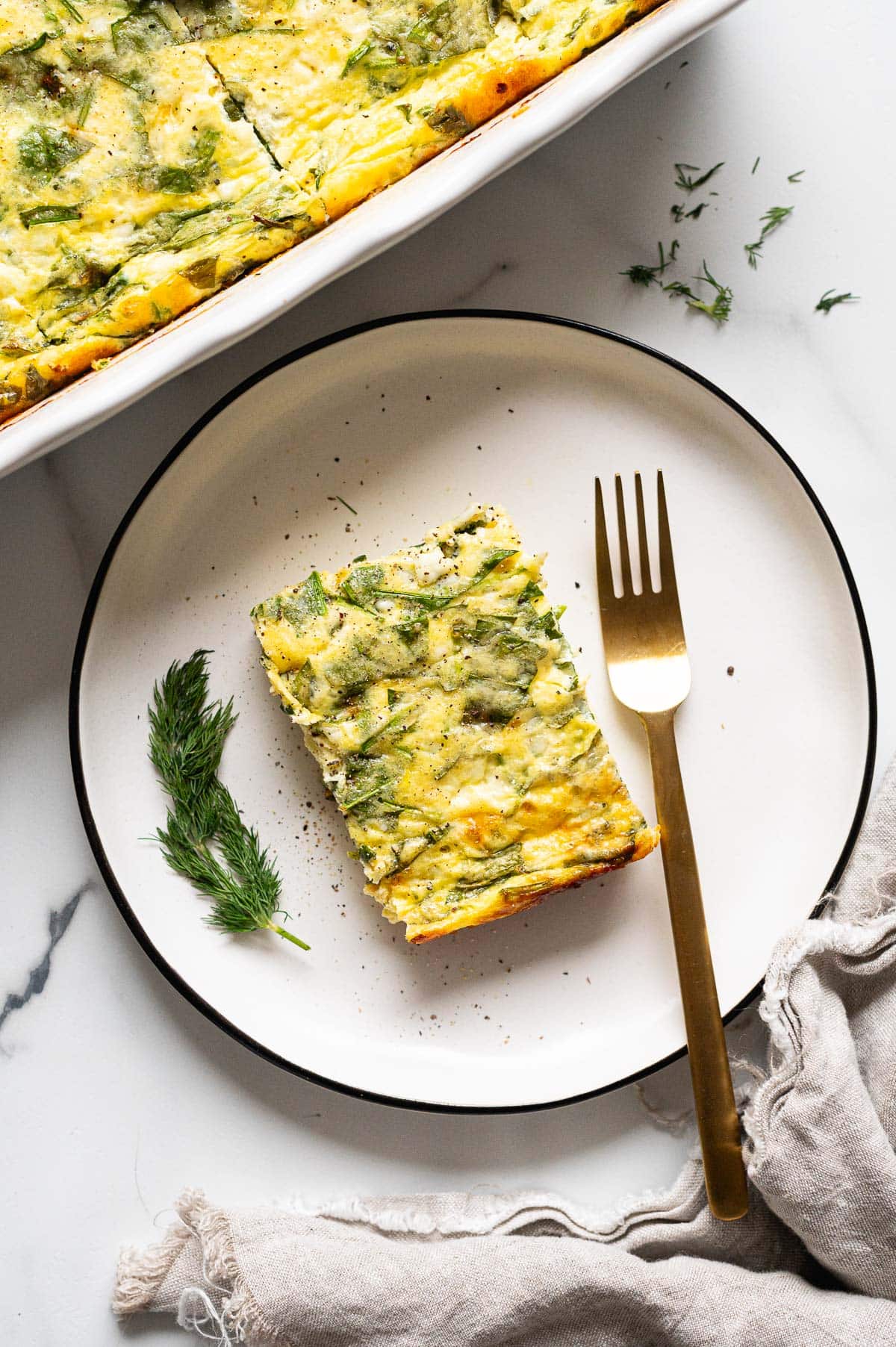Slice of egg bake on a plate with a fork and dill. Casserole in a dish next to it.