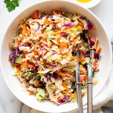 Easy coleslaw recipe served in a bowl with tongs. Oil, parsley and linen towel on the counter.