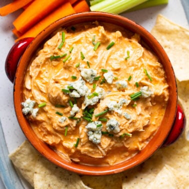 Instant pot buffalo chicken dip served in a bowl with blue cheese crumbles and chives. Chips and vegetables on a platter.