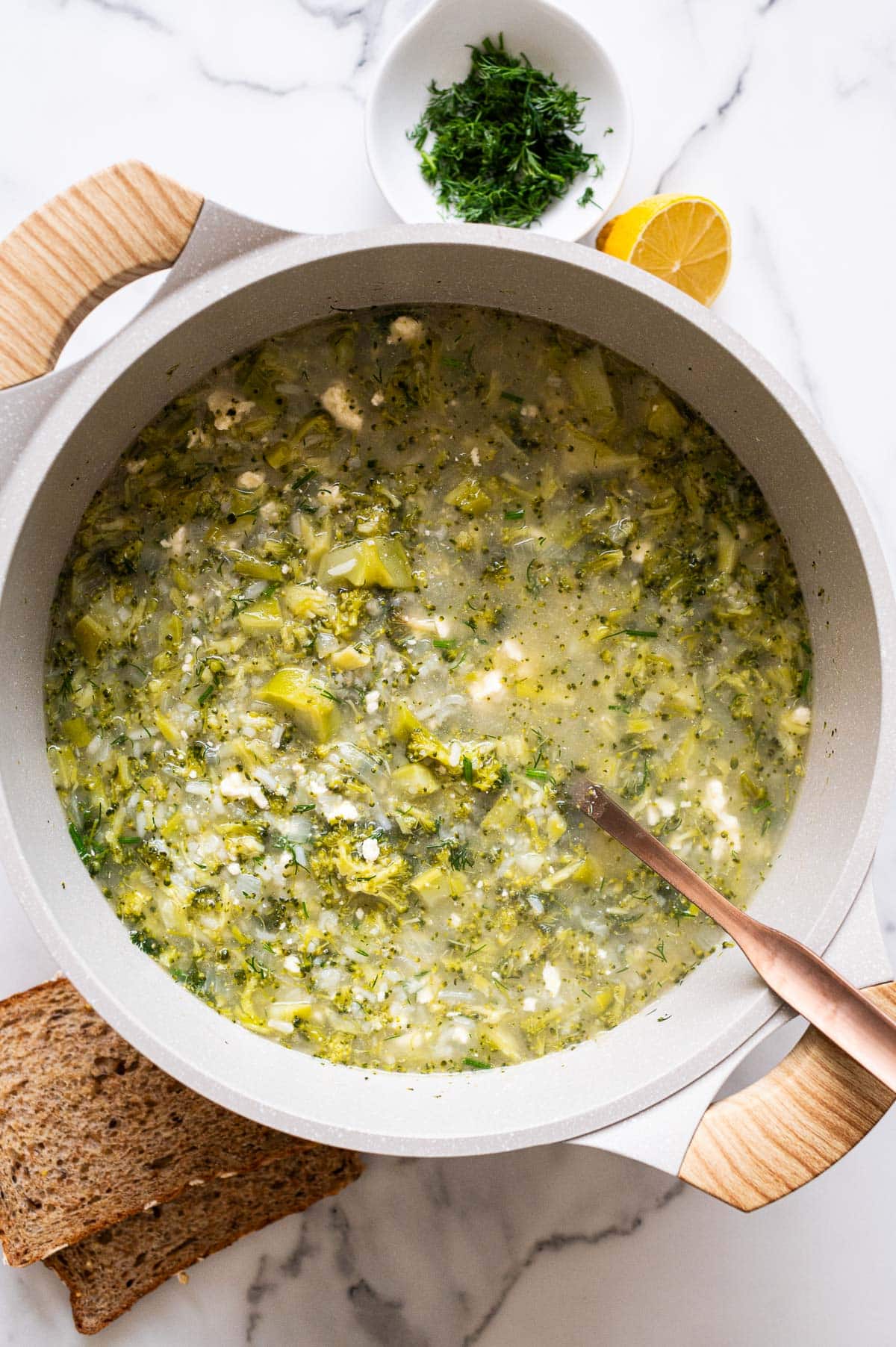 Broccoli feta soup in a pot with ladle. Dill, lemon and bread on a counter.