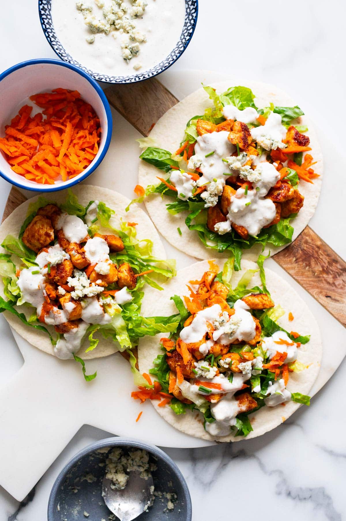 Buffalo chicken tacos drizzled with blue cheese dressing on a marble board. Shredded carrots and dressing in bowls.