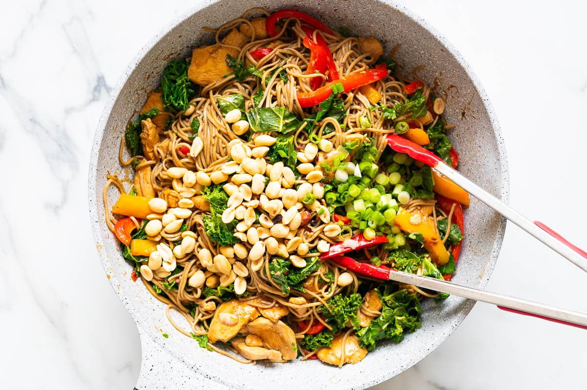 Soba noodles, peanuts, chicken, bell peppers, kale and green onion in a skillet with tongs.