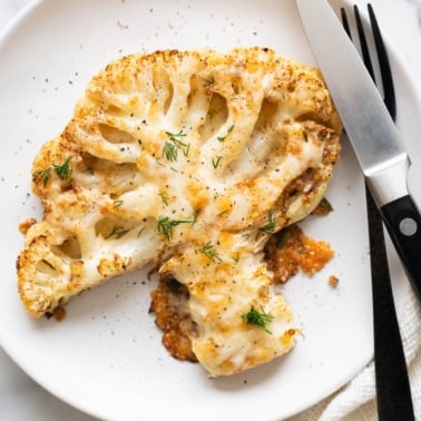 Cheesy cauliflower steak garnished with dill and served on a plate with utensils.