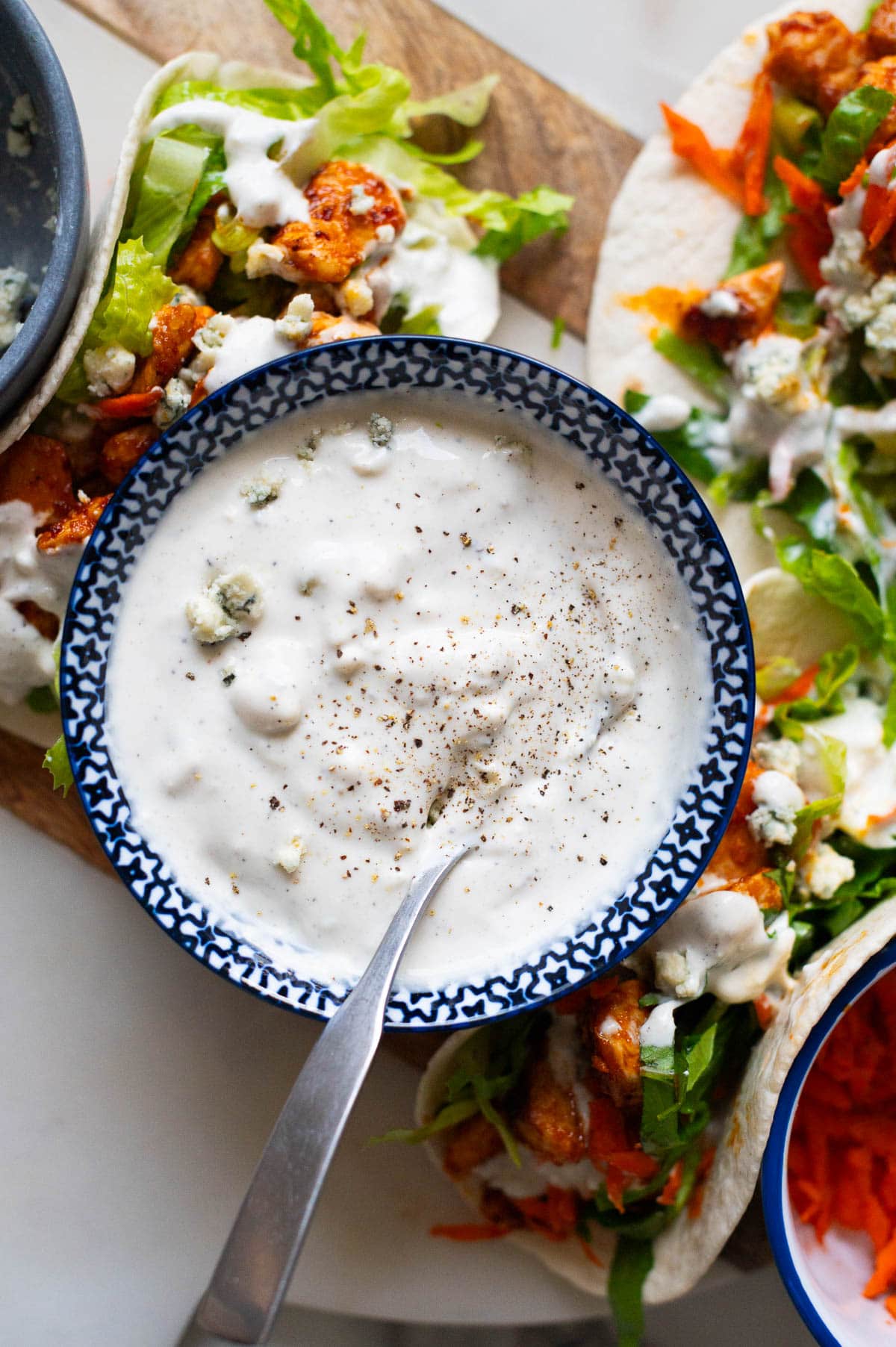 Greek yogurt blue cheese dressing in a bowl with a spoon. Buffalo chicken tacos around it on a board.