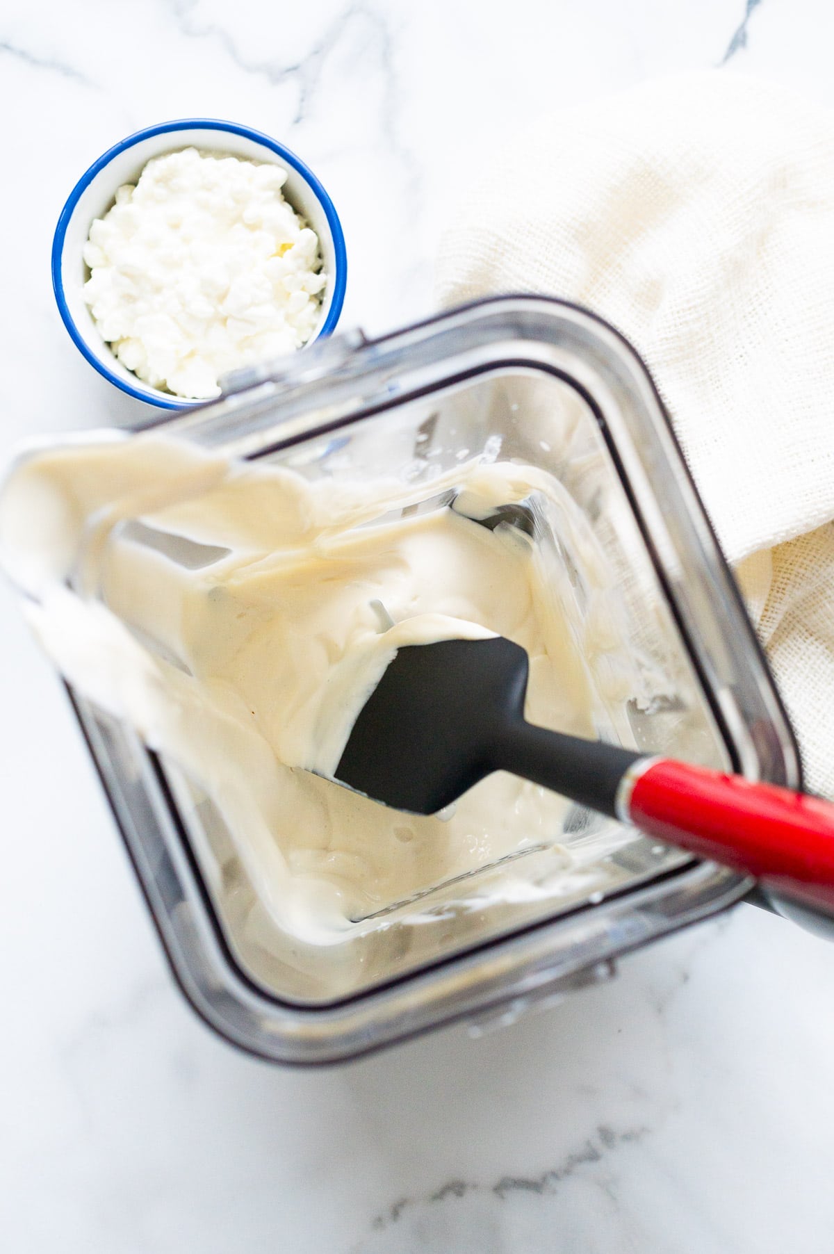 Cottage cheese protein pudding in a blender with spatula. A bowl with cottage cheese and linen napkin on a counter.