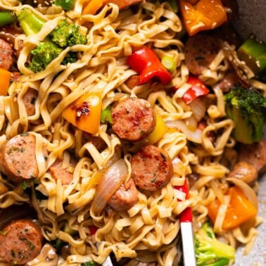 Sausage stir fry with broccoli, onions, bell peppers and noodles in a wok with tongs.