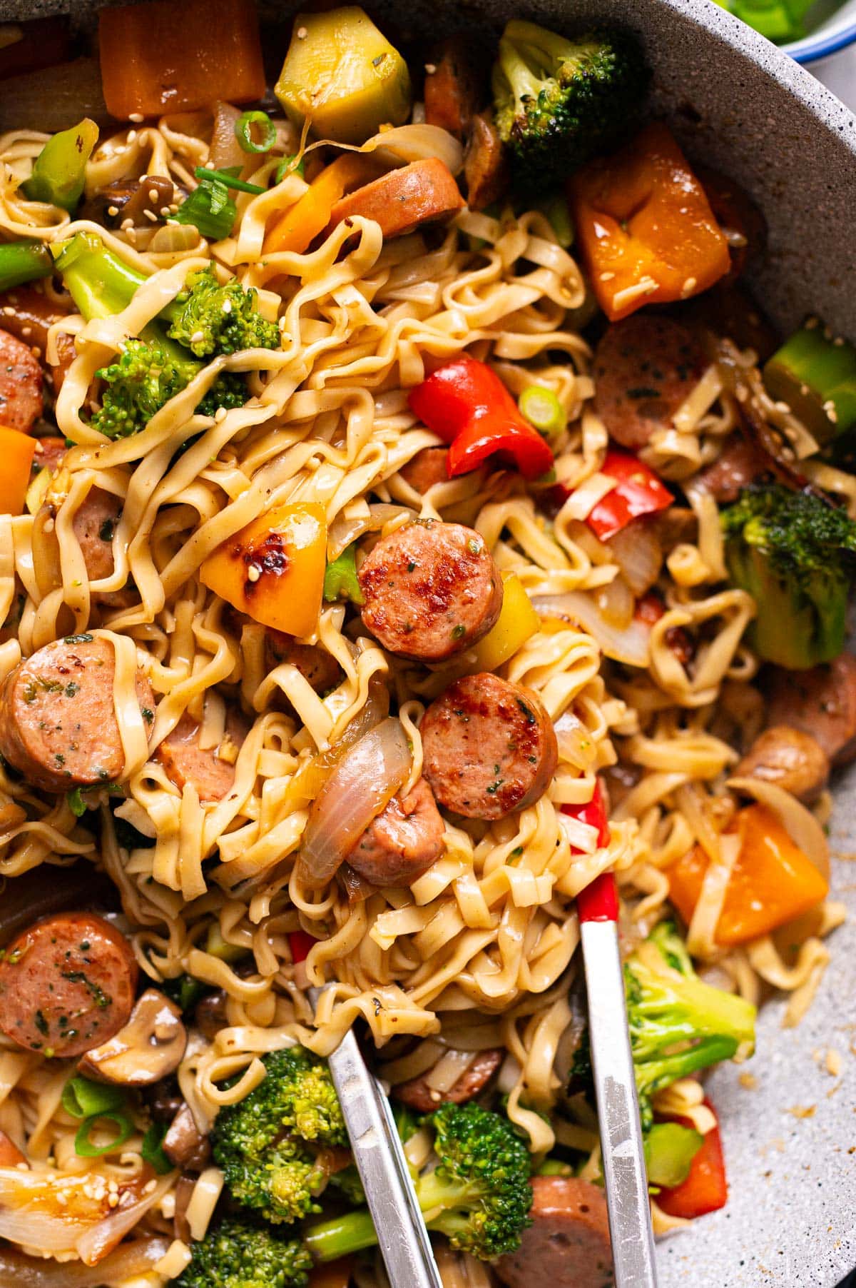 Sausage stir fry with broccoli, onions, bell peppers and noodles in a wok with tongs.