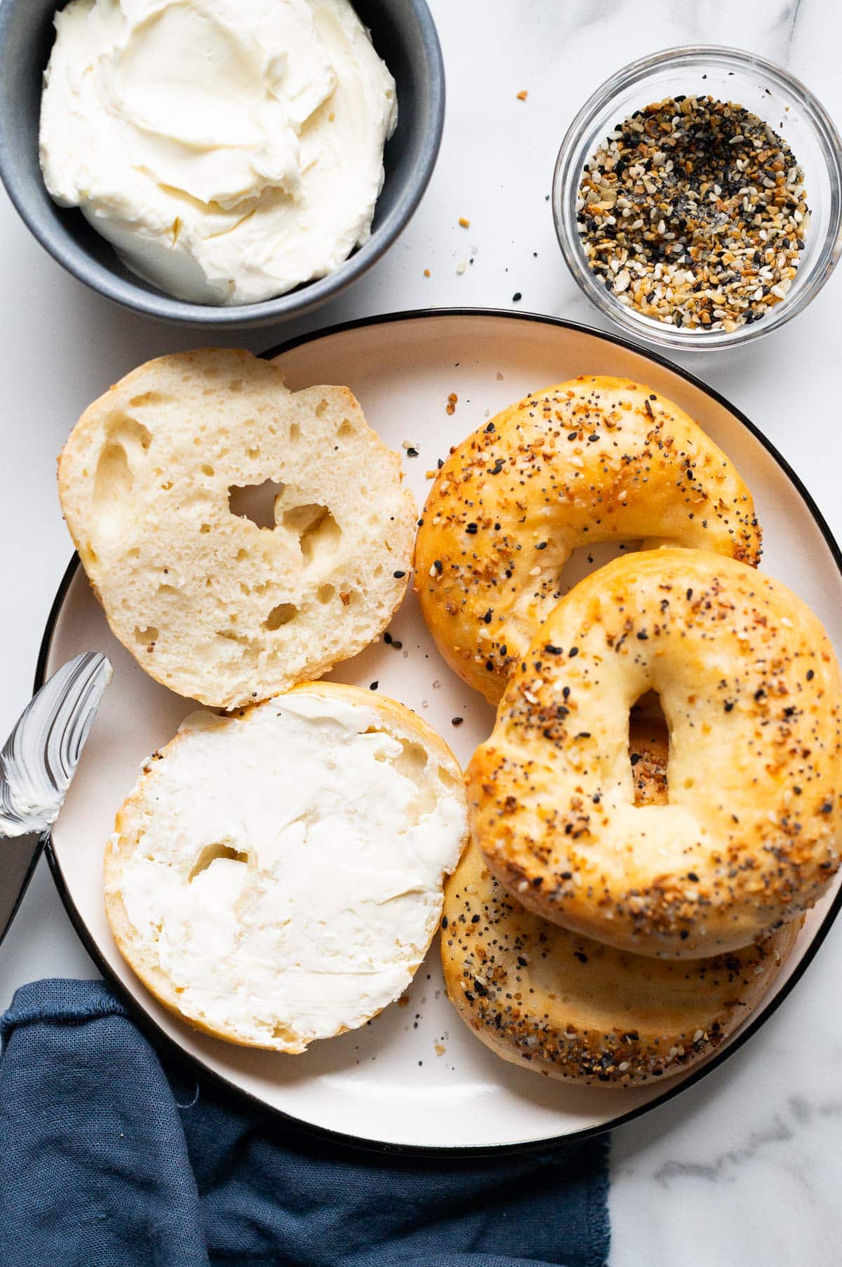 4 Greek yogurt Bagels on a plate 1 sliced and spread with cream cheese. Cream cheese, everything bagel seasoning, knife and napkin on a counter.