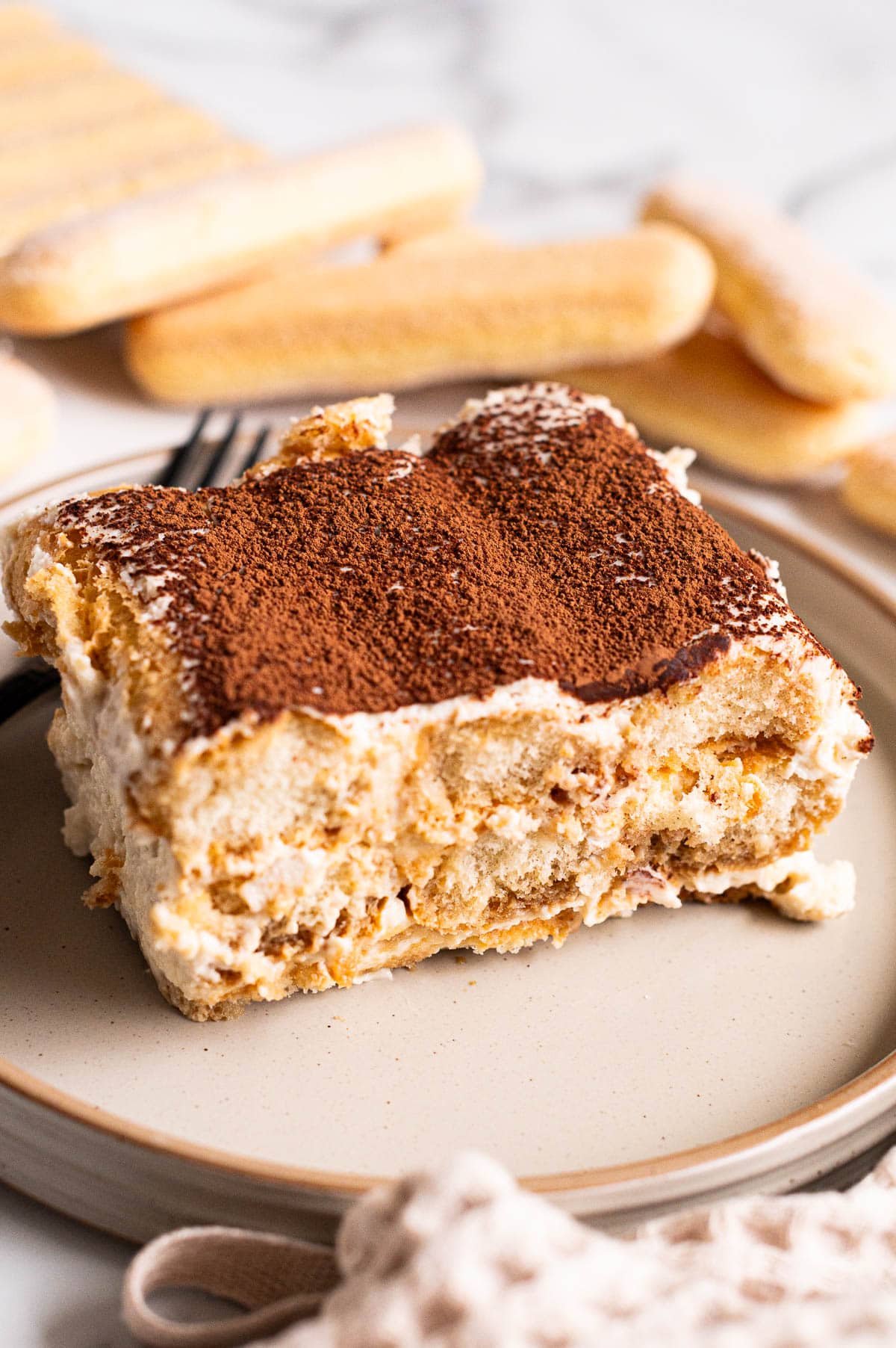 A slice of cottage cheese tiramisu served on a plate. Lady fingers on the counter.