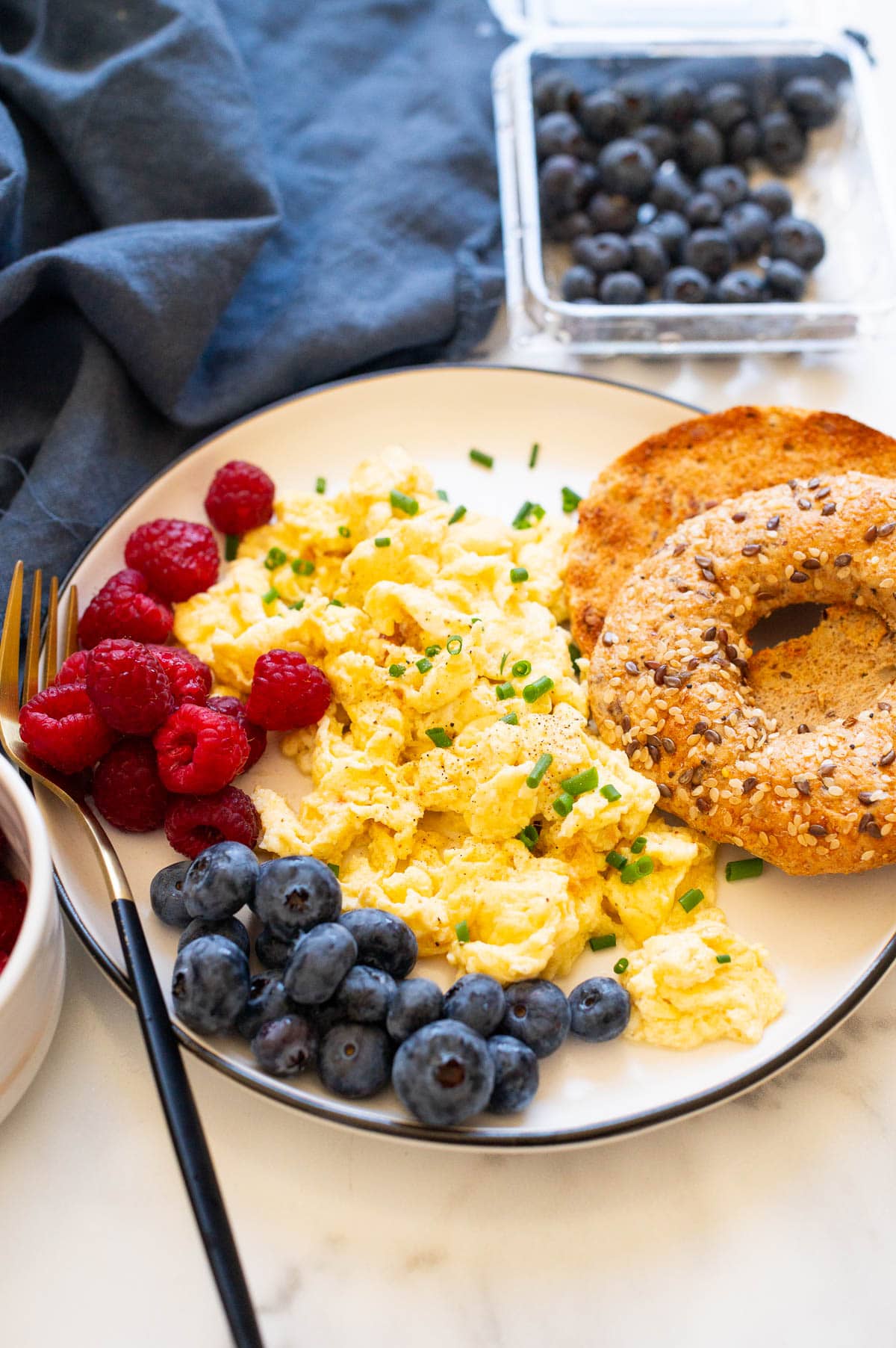 Side view of scrambled eggs garnished with chives and served on a plate with berries and toast. Napkin and berries in a container on a counter.