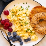 Cottage cheese scrambled eggs served on a plate with raspberries, blueberries and toasted bagel.