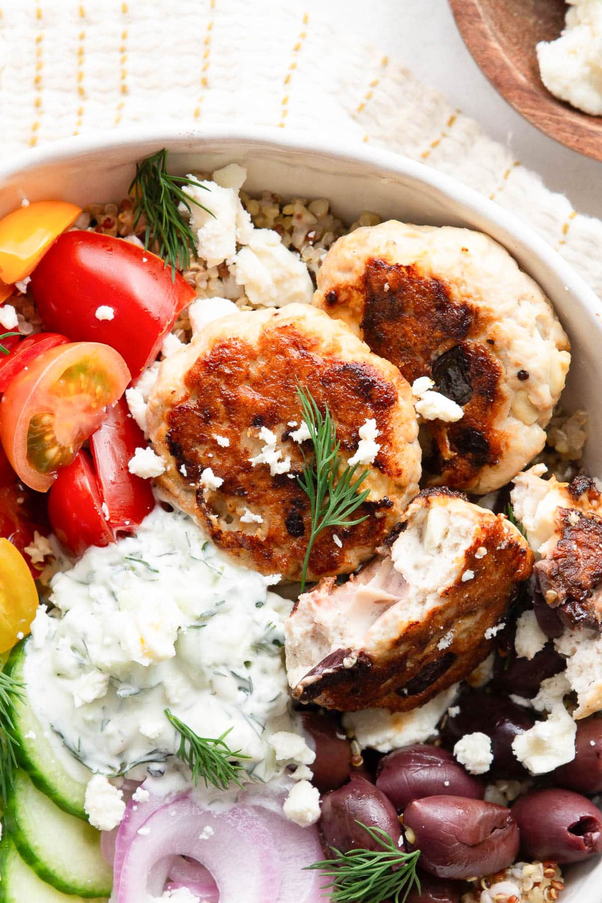 Close up of turkey burgers garnished with feta cheese, dill and tzatziki. Served in a bowl over quinoa with vegetables and olives.