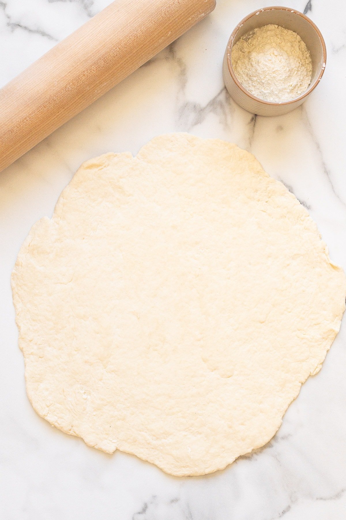 Rolled out pizza dough on the counter, rolling pin and flour in a bowl.