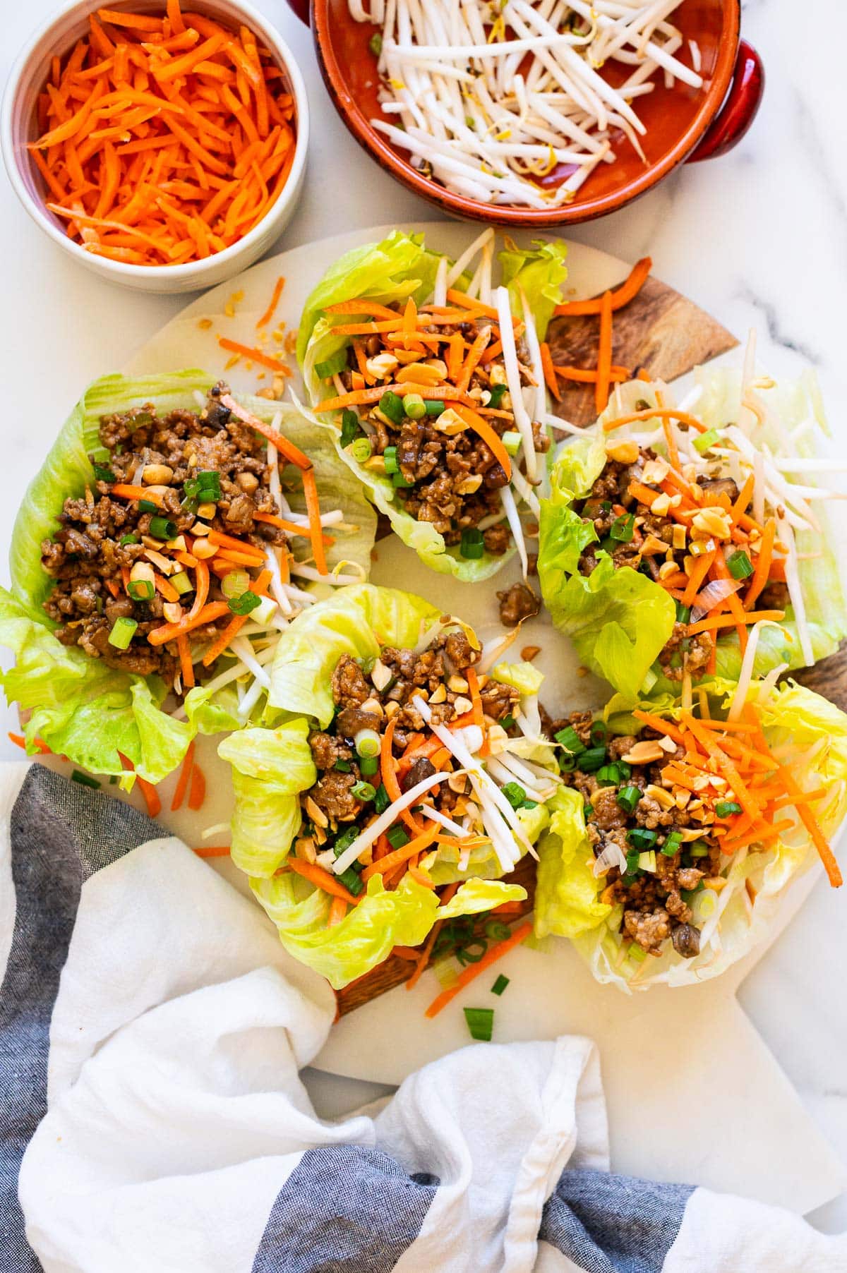 Looking down on a platter with five turkey lettuce wraps. Shredded carrots and bean sprouts in bowls. Linen towel on a counter.