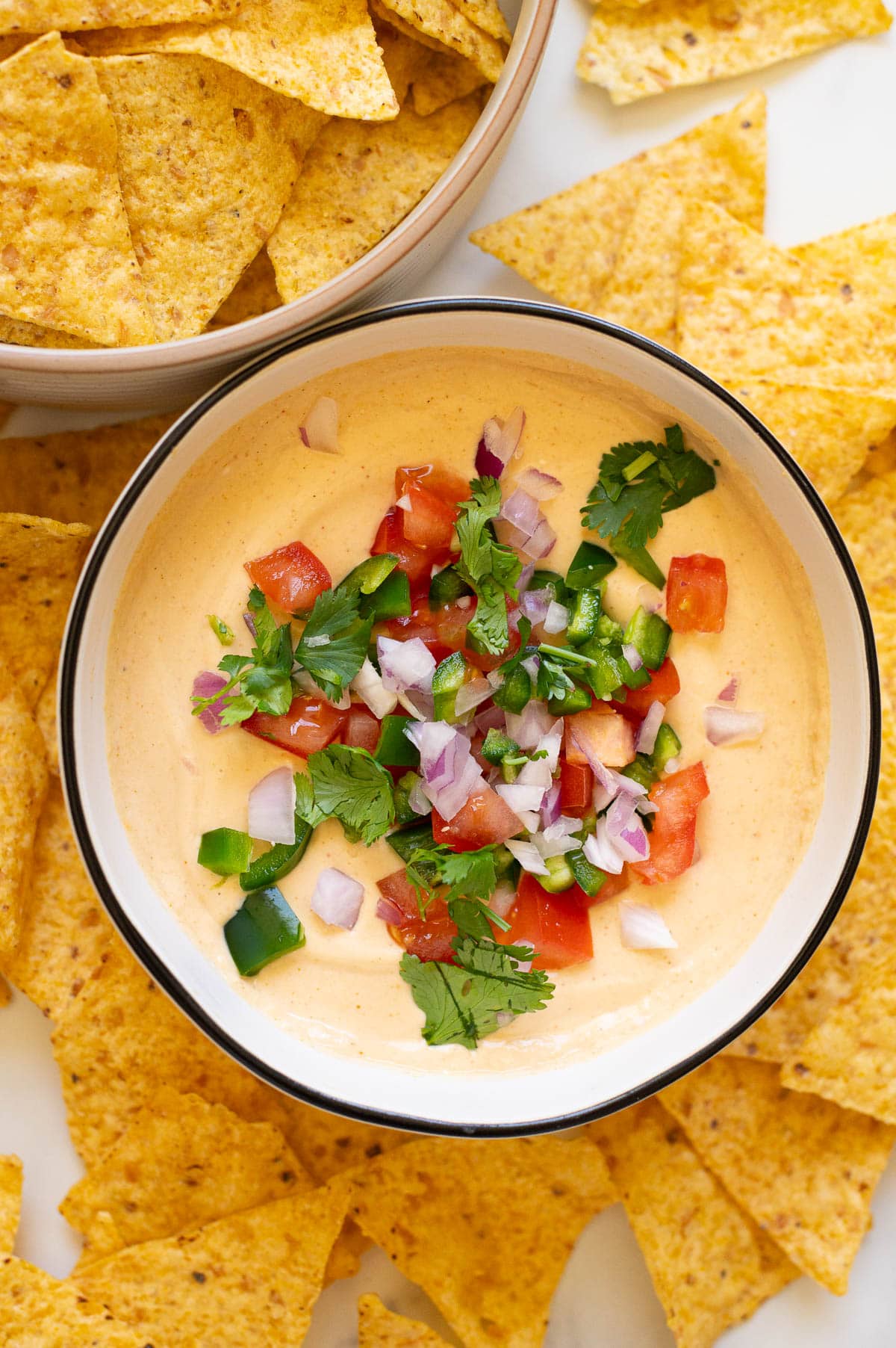 Cottage cheese queso dip in a bowl garnished with toppings. Tortilla chips in a bowl and on a counter around it.