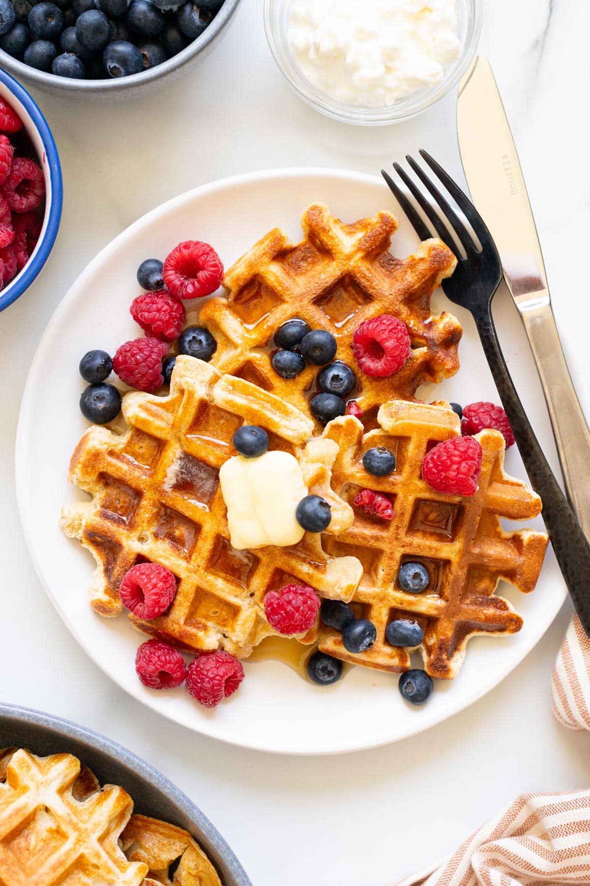 Cottage cheese waffles with berries, butter and maple syrup on a plate with a fork and knife. Toppings on a counter.