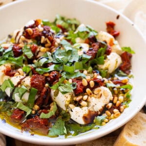 Burrata dip with sun-dried tomatoes, pine nuts and basil in a bowl. Bread slices around it.