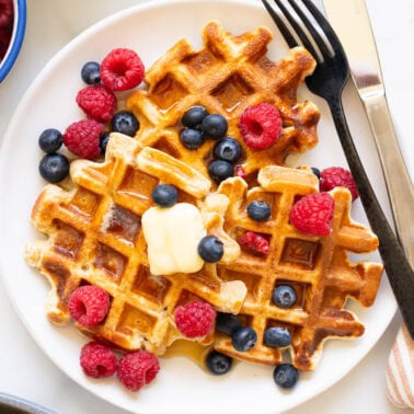 Cottage cheese waffles with berries, butter and maple syrup on a plate with a fork and knife.