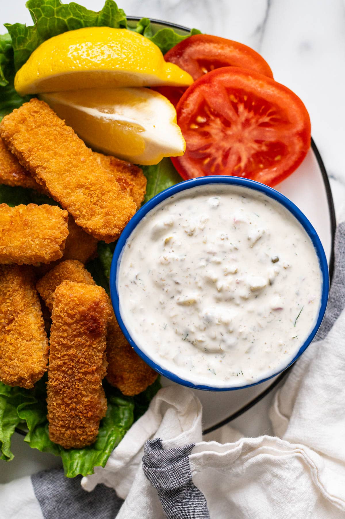 Tartar sauce in a bowl. Fish sticks, lemon wedges and tomato slices on a plate.