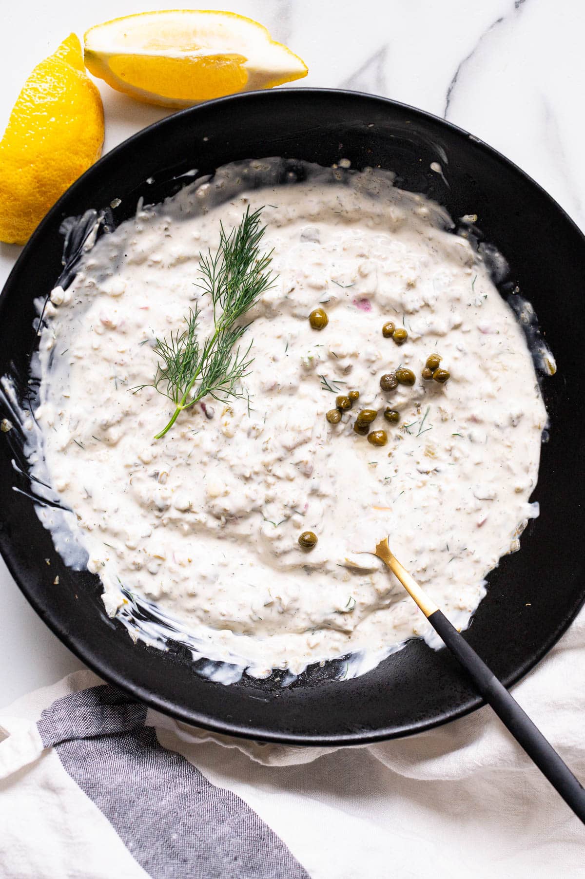 Greek yogurt tartar sauce  with capers and fresh dill in a black bowl with fork. Lemon wedges and towel on a counter.