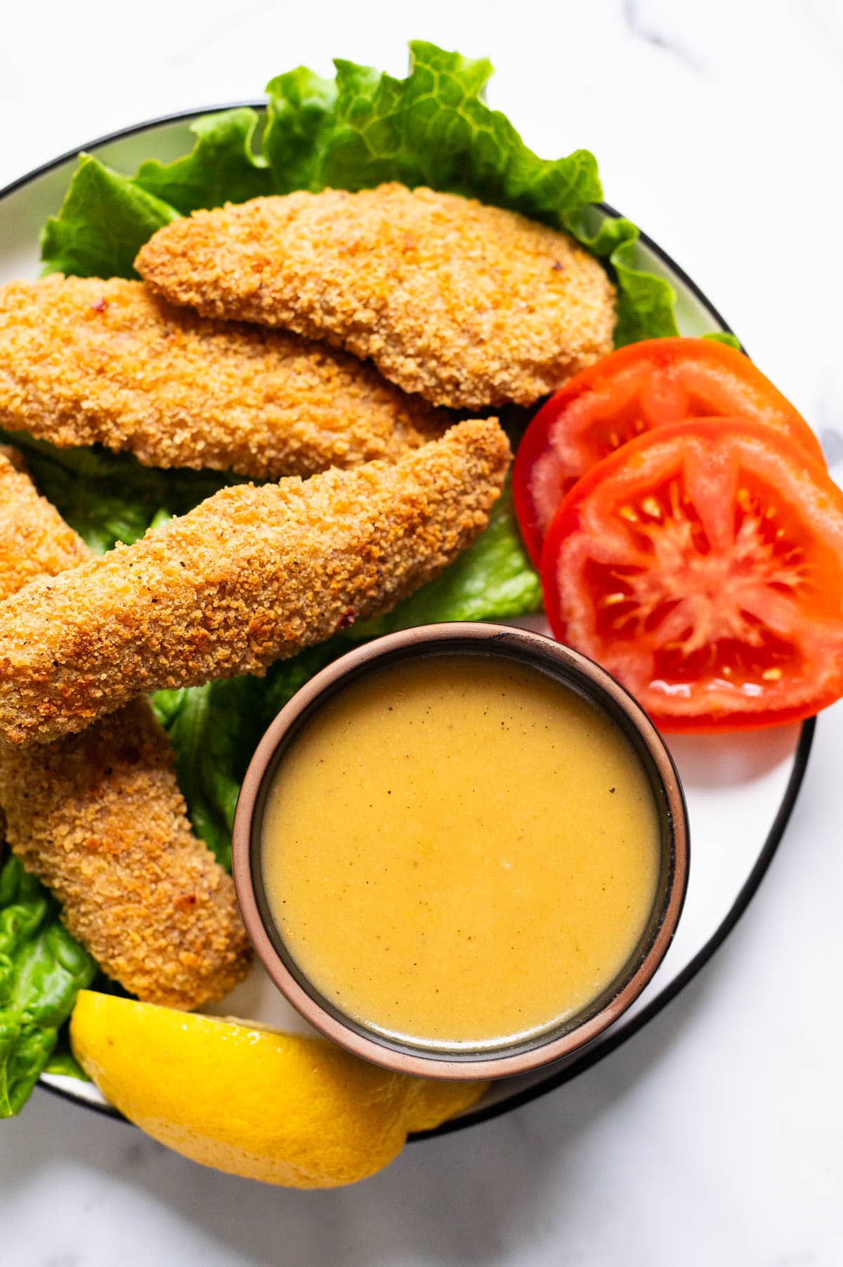 Healthy honey mustard dressing in a bowl with  chicken fingers and veggies on a plate.