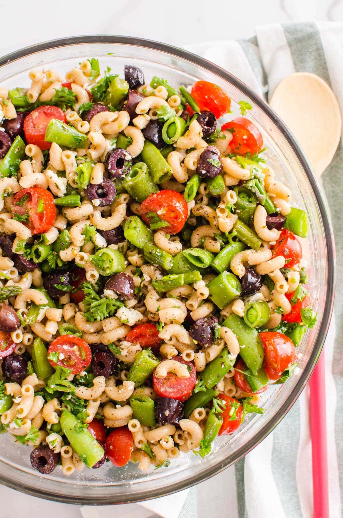 Looking down on a bowl of pasta salad with tomatoes, peas, olives and feta. Linen towel and wooden spoon on a counter.