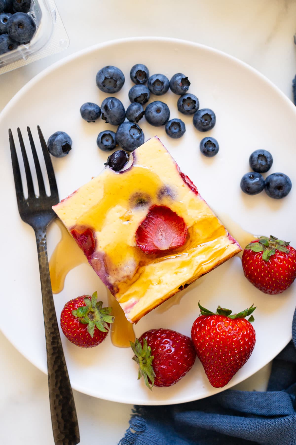 A slice of baked yogurt served with maple syrup and berries on a plate with a fork.