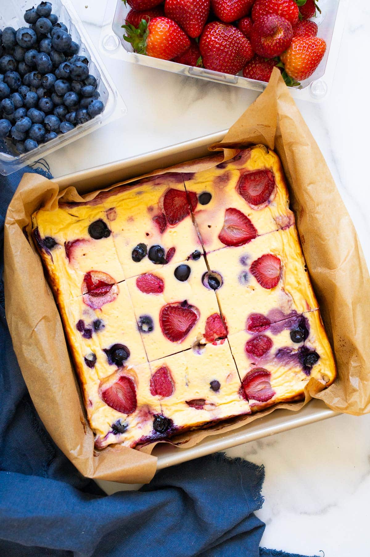 Baked yogurt with berries cut into nine slices in a baking dish. Blueberries, strawberries and napkin on a counter.