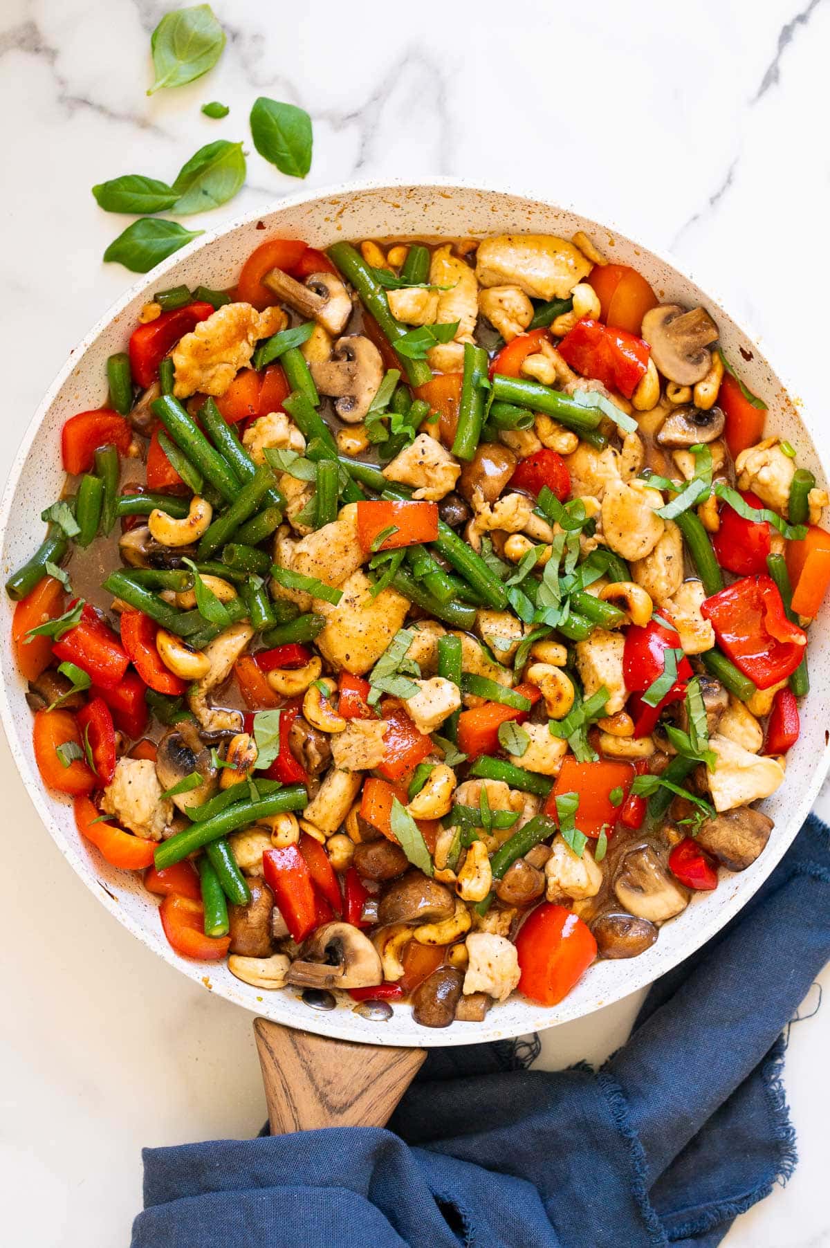 Basil cashew chicken with vegetables in a skillet. Blue napkin and basil leaves on a counter.