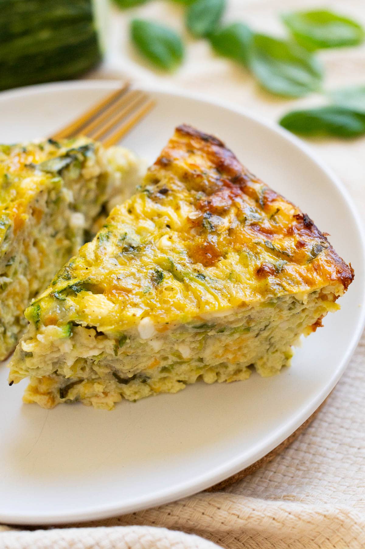 Two slices of crustless zucchini quiche on a plate with a fork. Fresh basil and zucchini in the background.