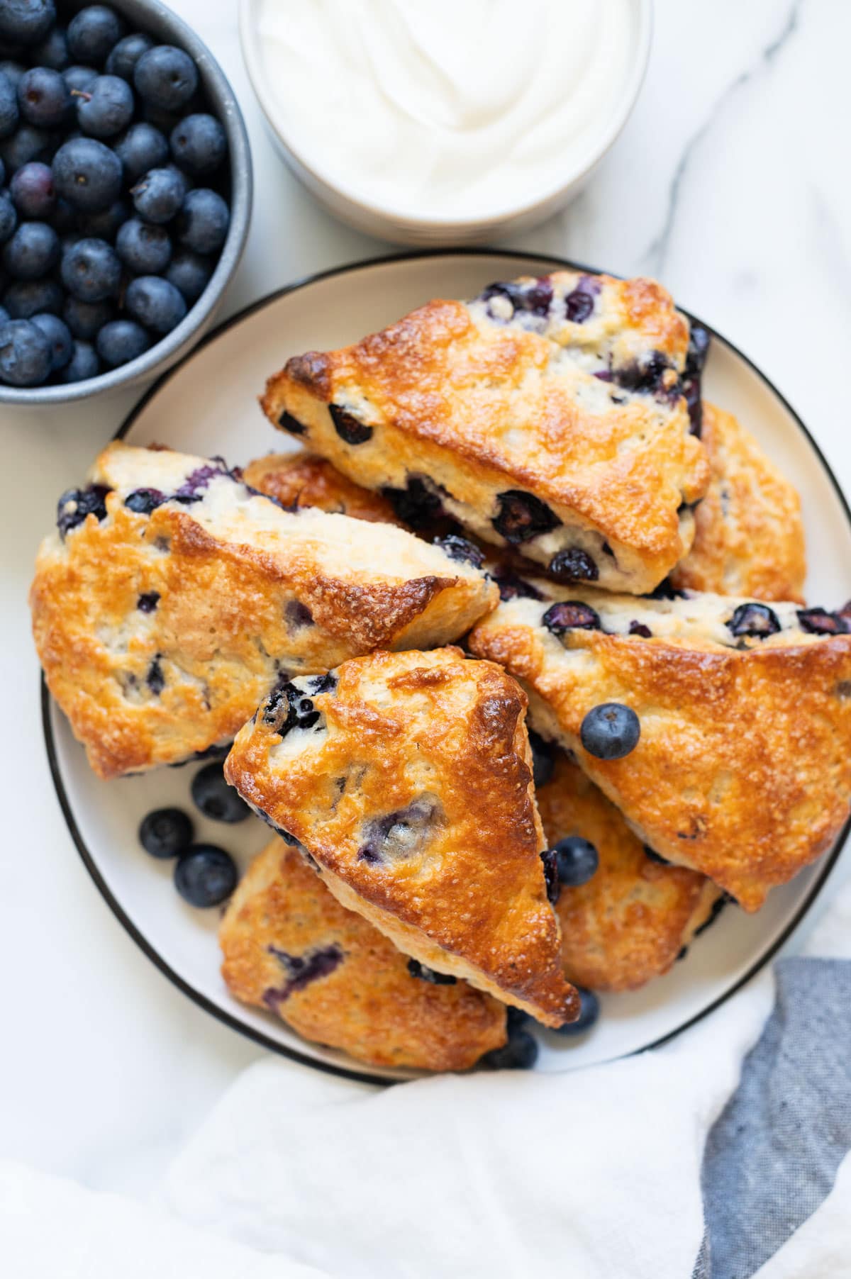 Blueberry scones on a plate. A bowl with blueberries and Greek yogurt and linen towel on a counter.