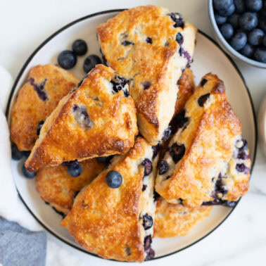 Blueberry scones on a plate. A bowl with blueberries and Greek yogurt and linen towel on a counter.