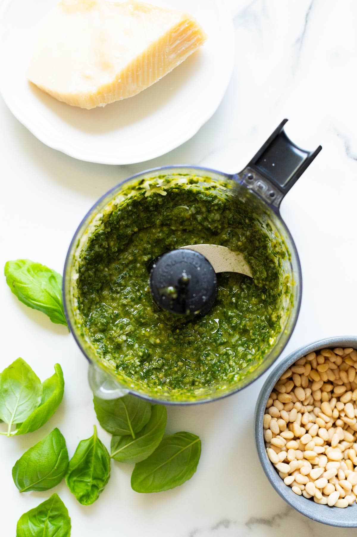 Basil pesto sauce in a food processor bowl. Pine nuts, basil and parmesan cheese on the countertop.