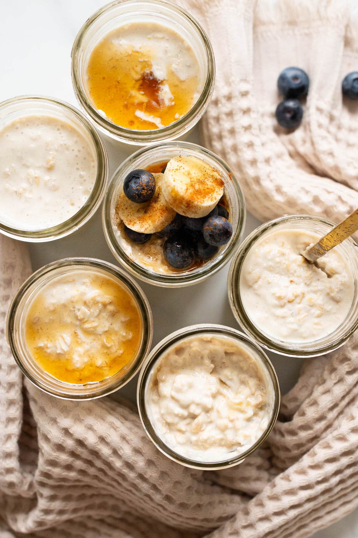 Six glass jars with overnight oats and topped with maple syrup and fruit. Towel on a countertop 