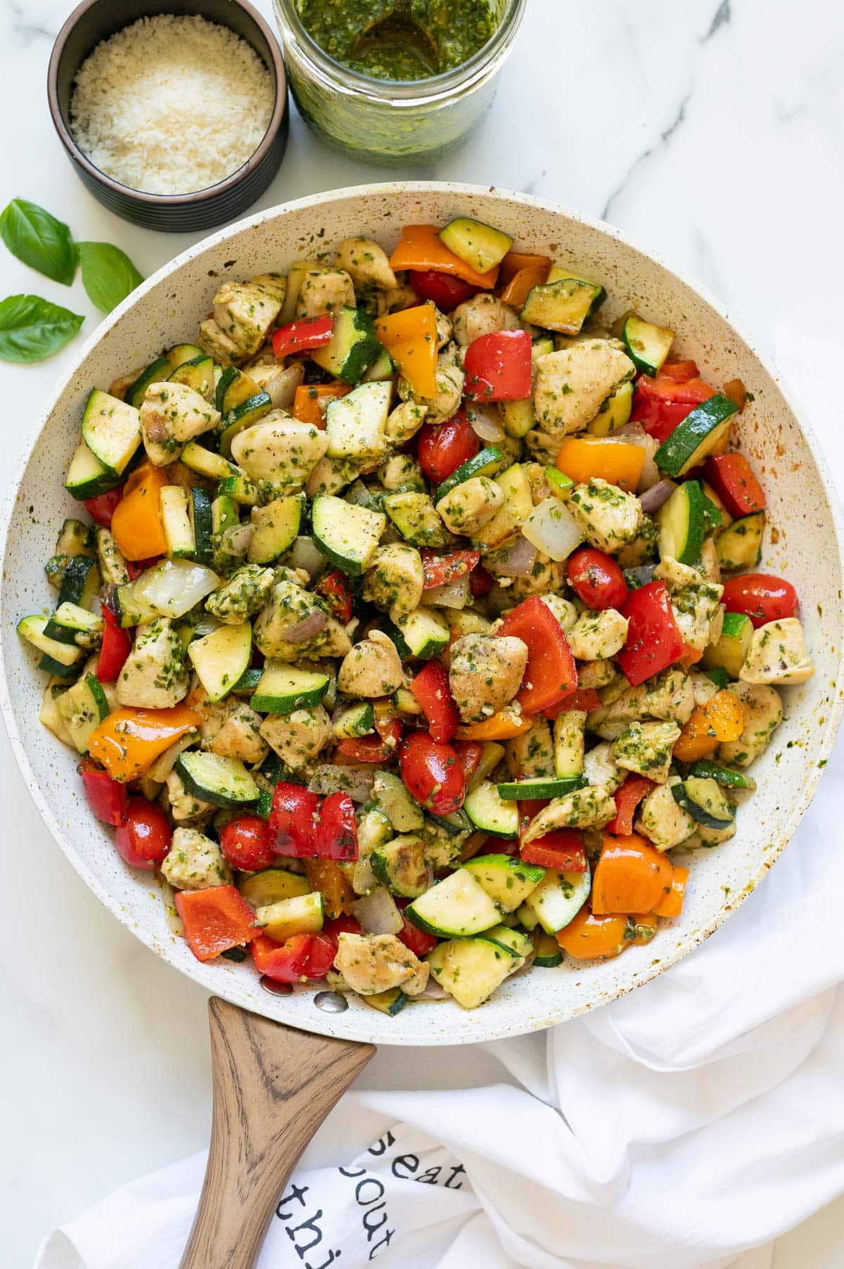 Pesto chicken and vegetables in a skillet. Pesto, parmesan cheese, basil and napkin on a counter.