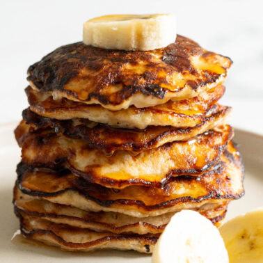 A stack of banana egg pancakes garnished with banana slices and drizzled with maple syrup on a plate.