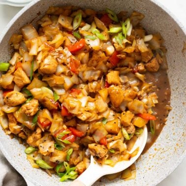 Chicken cabbage stir fry garnished with green onions in a skillet with a spoon.