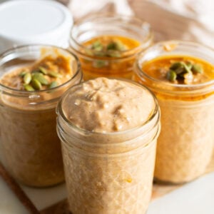 Pumpkin protein overnight oats topped with pumpkin seeds and maple syrup in glass jars.