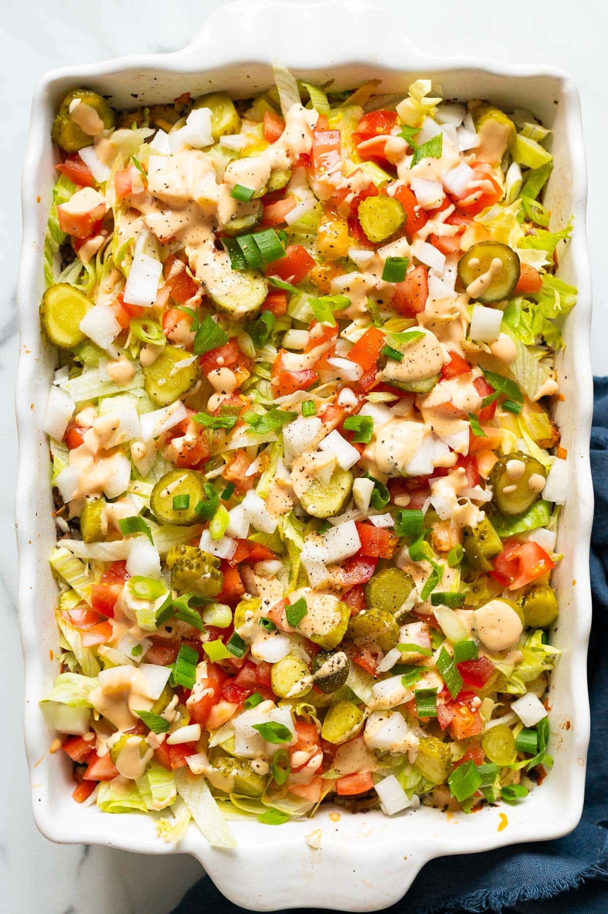 Big Mac casserole with sauce and toppings in a baking dish. Blue linen napkin on a counter.