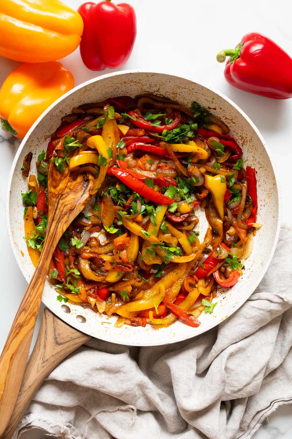 Sauteed peppers and onions in a skillet with a spoon. Bell peppers on the counter.