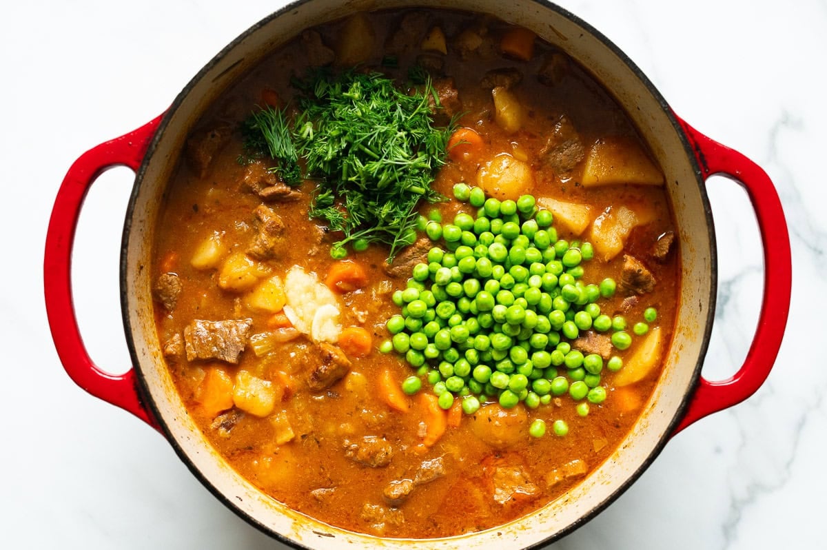 Dill, peas and garlic added to cooked beef stew in a pot.