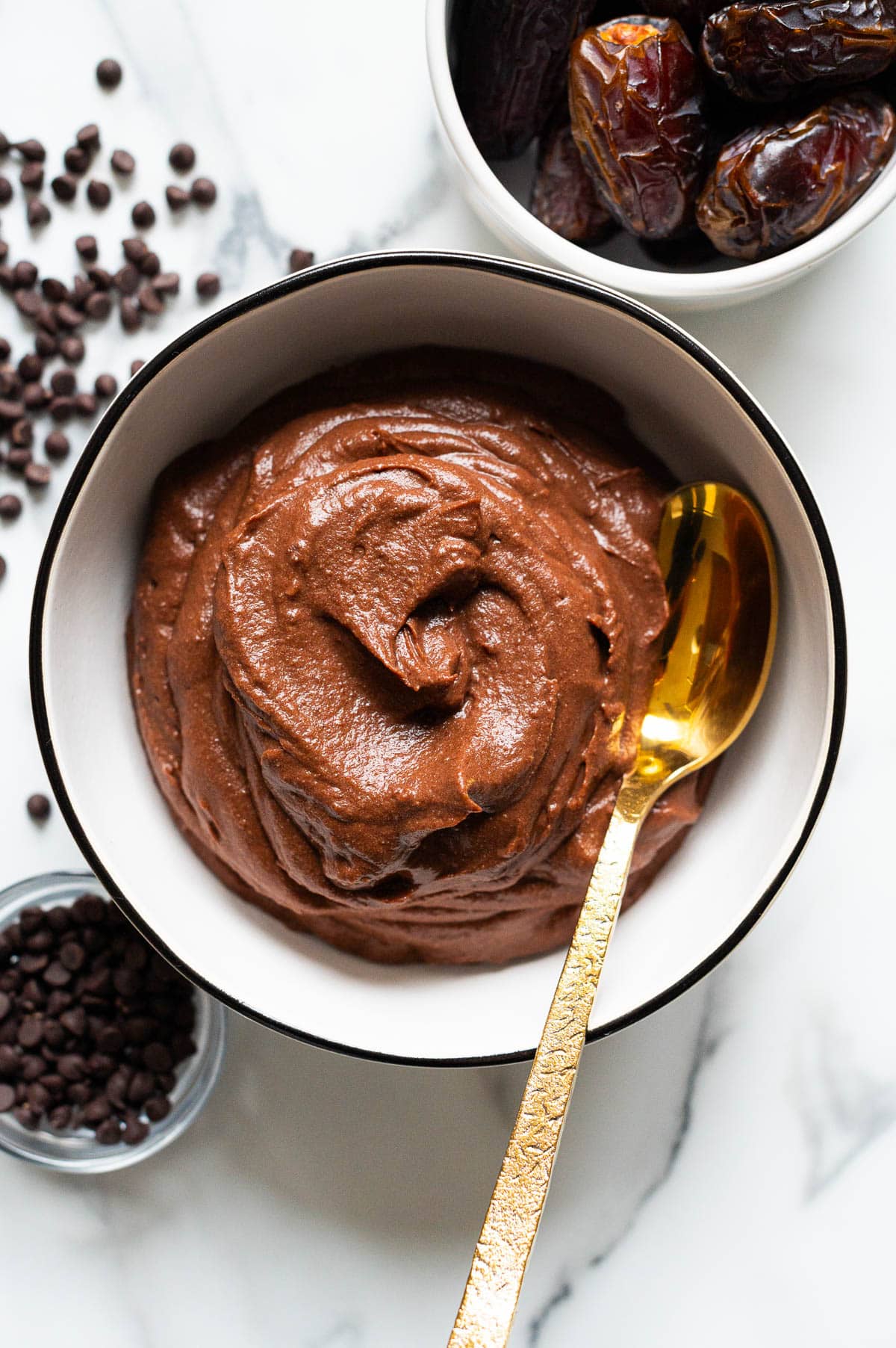 Hard boiled egg pudding in a bowl with a spoon. Bowl with dates and chocolate chips on a counter.