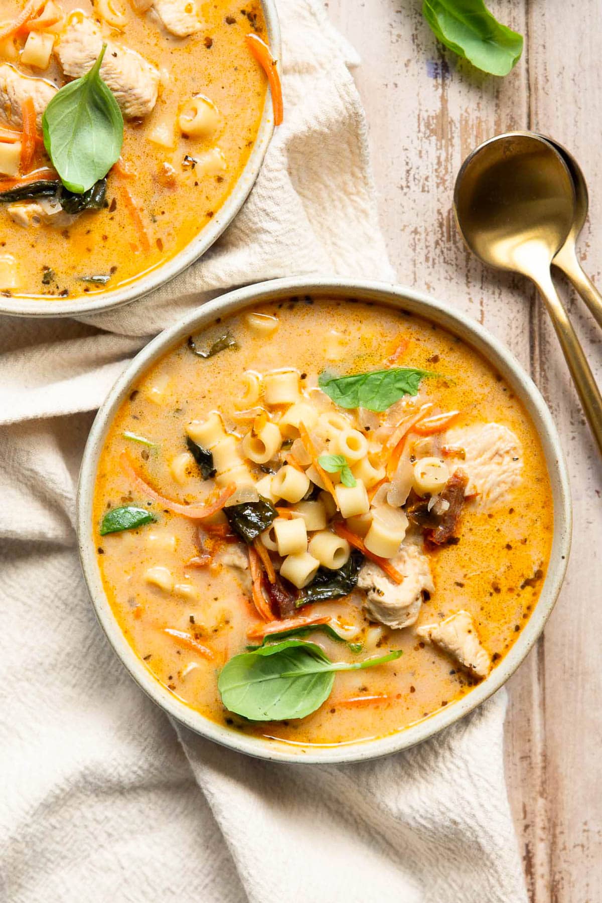 Two bowls with marry me chicken soup. Spoons, napkin and basil on a counter.